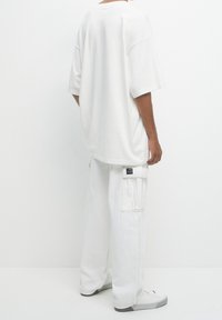 T-shirt blanc surdimensionné à manches courtes, associé à un pantalon cargo blanc avec poches latérales et une étiquette à l'arrière. Baskets blanches incluses.