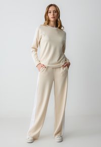 Beige loungewear set met een langwerpige top met witte accenten en losse broek met zijzakken. Zachte, gladde textuur.