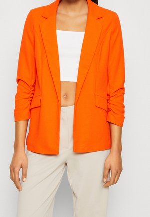 Kobieta ubrana w jasnopomarańczową marynarkę na biały crop top i jasnobeżowe spodnie z wysokim stanem na jasnym tle.