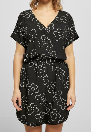 Žena v černých šatech s velkými bílými květinovými vzory, krátkými rukávy, výstřihem do V a páskem v pase.