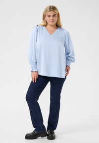 Blouse bleu clair à col en V avec des manches bouffantes et des poignets élastiques, associée à un jean bleu foncé et des chaussures à plateforme noires. Design simple et fluide.
