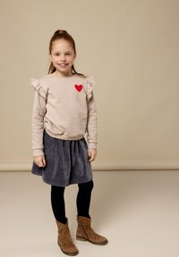 Beige sweater met franje op de schouders en een rood hartontwerp, gecombineerd met een grijze corduroy rok, zwarte maillots en bruine enkellaarsjes.