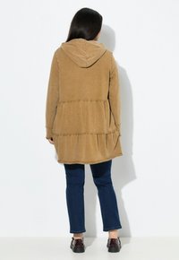 Indumento marrone con cappuccio, a maniche lunghe e dettagli con orlo arricciato; indossato sopra jeans scuri e scarpe nere. Texture morbida, design casual.