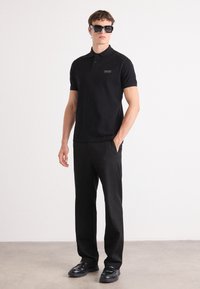 Polo noir à manches courtes avec un logo de poche, assorti à un pantalon noir et des mocassins noirs brillants. Modèle portant de grandes lunettes de soleil noires.