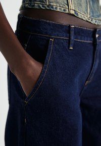 Tmavě modré džíny z denimu s žlutým prošíváním, předními kapsami a přiléhavým střihem viditelným na bocích.