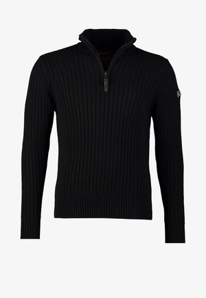 Homme marchant portant un pull à fermeture éclair noir, un jean bleu, des bottes marron et une bandoulière de sac marron sur l'épaule.