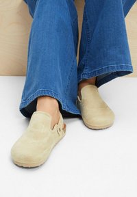 Sabots en suède beige clair avec un design sans arrière, une semelle solide et une ouverture latérale. Portés avec un pantalon en denim bleu évasé.