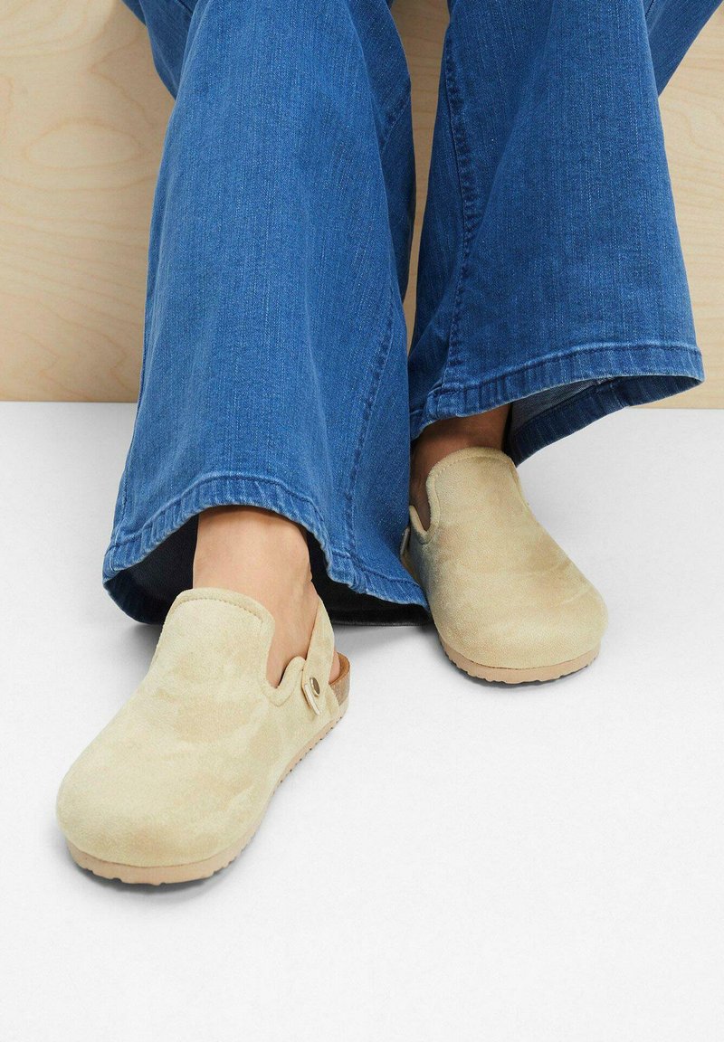 Sabots en suède beige clair avec un design sans arrière, une semelle solide et une ouverture latérale. Portés avec un pantalon en denim bleu évasé.