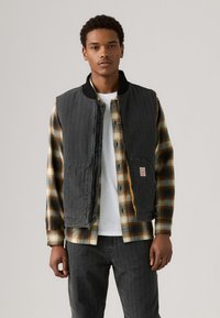 Črn prešit telovnik preko rjave in rumene karo flanelaste srajce. Komplet vsebuje belo majico pod njim in temno denim hlače.