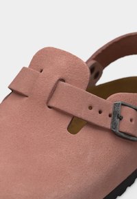 Κοντινό πλάνο ενός ροζ σουέντ τσόγκου με φαρδιά στεφάνη και μεταλλικό κουμπί πάνω από τον καρπό του ποδιού και μαύρη υφασμάτινη σόλα.