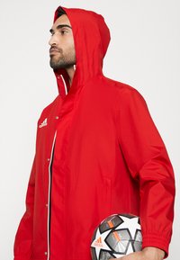 Rode waterdichte jas met een ritssluiting aan de voorkant, elastische manchetten en een capuchon. Kenmerkt zich door witte accenten en een Adidas-logo. Houdt een zwart-witte voetbal vast.