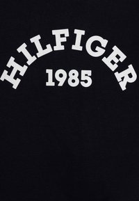 Tela negra con texto curvado en blanco "HILFIGER 1985" mostrado de forma destacada; textura suave, material de peso medio, sin patrones ni adornos.