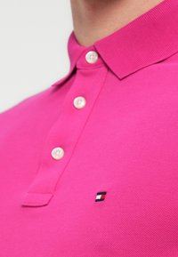 Ροζ πόλο πουκάμισο από υφή ύφασμα, με τρεις κουμπότρυπες και μικρό κεντημένο λογότυπο σε μπλε, κόκκινο και λευκό.