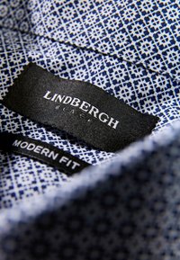 Marineblauw stof met een geometrisch patroon, met zwarte labels waarop "LINDBERGH BLACK" en "MODERN FIT" staan. Close-up van de kraag.