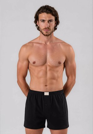 Muscular man with short dark hair wearing black boxer shorts stands against plain gray background, hands behind his back.
