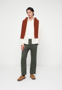 Férfi áll, bézs inget visel fehér póló felett, rozsdabarna pulóver a vállán, olívazöld nadrágot és világosbarna velúr cipőt visel.