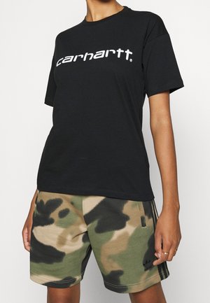 Osoba ubrana w czarną koszulkę Carhartt i zielone, beżowe oraz czarne krótkie spodenki Adidas w kamuflażu, stojąca z rozluźnionymi ramionami.