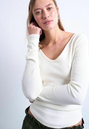 Young woman with light brown hair wearing a white ribbed long-sleeve top and dark green pants, posing with one hand near her face.