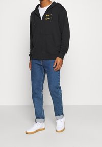 Black zip-up hoodie with a gold Nike logo, paired with blue jeans and white sneakers. The fabric appears soft with a relaxed fit.