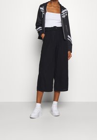 Svarta korta byxor med veck, hög midja och bälte. Kombineras med en vit linne och en svart jacka med dragkedja och grå ränder. Vita sneakers.