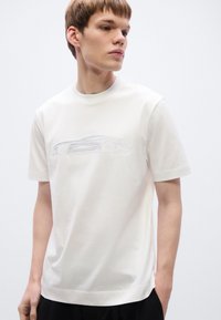 Άσπρο βαμβακερό T-shirt με λεπτό ανάγλυφο σχέδιο σιλουέτας αυτοκινήτου στο μπροστινό μέρος. Κοντά μανίκια και στρογγυλή λαιμόκοψη.