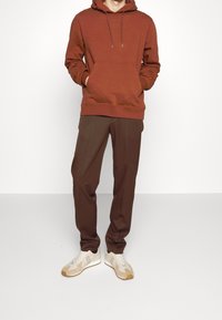 Sweat à capuche marron avec une poche avant, associé à un pantalon marron foncé et des baskets beige. La tenue présente une coupe décontractée et une texture douce.
