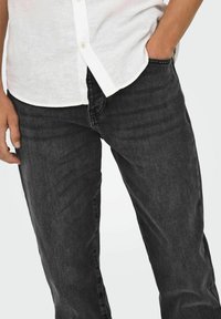 Jeans de mezclilla negra con corte recto, textura sutil y diseño clásico de cinco bolsillos, combinados con una camisa blanca de botones.
