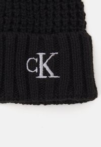 Črna pletena kapa z rebrasto teksturo, ki ima na sprednji strani belo izvezeno "CK" logo. Mehka in raztegljiva oblika.