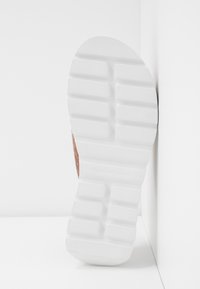 Semelle de chaussure blanche avec des rainures rectangulaires profondes en motif quadrillé, positionnée verticalement contre un fond d'angle blanc.