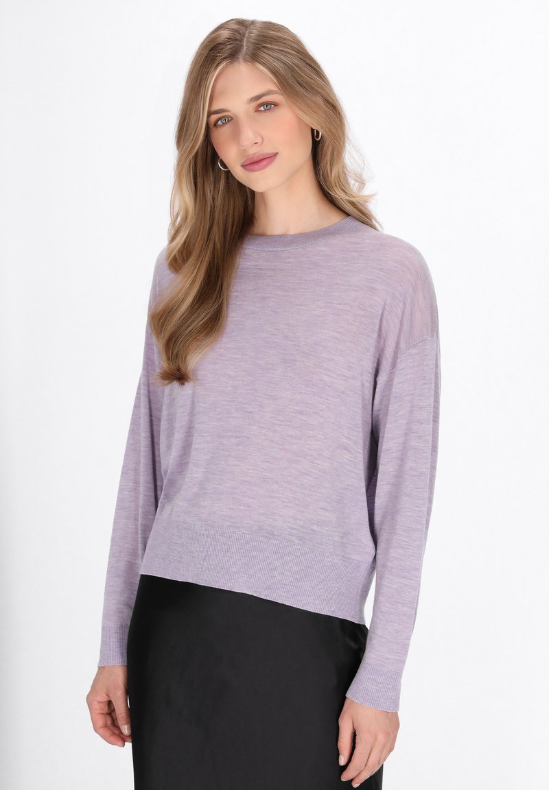 Maglione lungo a maniche con una vestibilità rilassata di colore viola chiaro, con polsini e orlo a coste, mostrato su una gonna nera contro uno sfondo neutro.