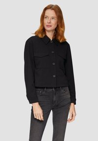 Vrouw met rood haar, gekleed in een zwarte blouse met knopen en voorzakken, en donkergrijze jeans, die lichtjes naar de zijkant kijkt.