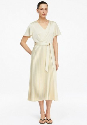 Kobieta stoi z ręką na biodrze, ubrana w kremową sukienkę z dekoltem w serek, krótkimi rękawami, plisowaną spódnicę midi, związany pasek w talii i czarne sandały.
