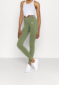 Leggings verdes de corte ajustado, con cintura alta y un sutil detalle de logo. Combinados con una camiseta sin mangas blanca atada y zapatillas blancas.