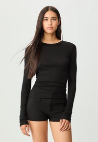 Schwarzes, langärmliges, geripptes Oberteil mit rundem Halsausschnitt, kombiniert mit schwarzen Shorts. Der Stoff wirkt weich und hat ein figurbetontes Design mit langen Ärmeln.