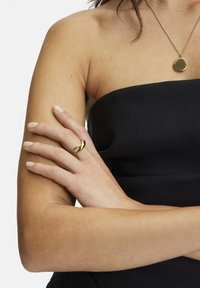 Anello d'oro con un design intrecciato indossato su una mano, abbinato a una collana con pendente d'oro coordinato. Top nero senza spalline visibile.