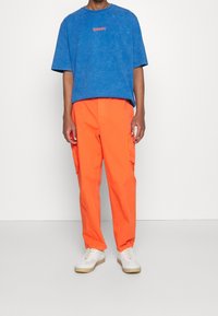 Blå överdimensionerad bomullströja med framlogotyp, matchad med ljusorange cargobyxor med sidofickor och vita sneakers med beige sulor.