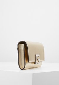 Sac à main métallique doré avec rabat à pli, doté d'une fermeture logo ornée de strass, texture lisse et forme structurée.