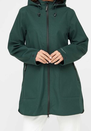 Kobieta ubrana w ciemnozielony płaszcz z kapturem z czarnym zamkiem błyskawicznym, bocznymi kieszeniami i regulowanymi zatrzaskami przy nadgarstkach, stojąca na jednolitym tle.