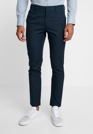 Homme portant un pantalon slim-fit bleu marine foncé, une chemise bleu clair boutonnée et des baskets blanches, debout devant un fond uni clair.