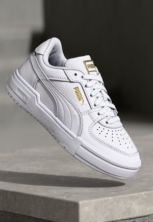 Sneaker bianco con punteggiature sulla punta, materiale in pelle e logo Puma dorato. Presenta pannelli laterali testurizzati e suola piatta in gomma.