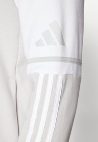 Grå og hvid atletisk sweatshirt med tre hvide lodrette striber på ærmet og et lys gråt logo på skulderen. Blødt stof.
