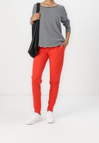 Gestreiftes Langarmshirt in Schwarz und Weiß, kombiniert mit engen roten Hosen und weißen Sneakers, hält eine schwarze Schultertasche.