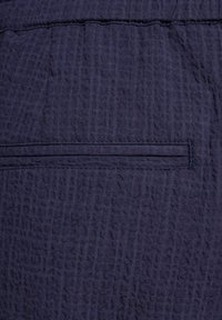 Tissu bleu marine texturé avec un motif froissé subtil. Présente une couture plate et des piqûres ainsi qu'une poche horizontale étroite à l'arrière.