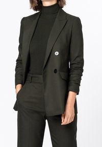Tmavě zelený upravený blazer s dvojitými knoflíky, zkombinovaný s riflovým rolákem. Hladká látka, strukturované ramena a přiléhavý silueta.