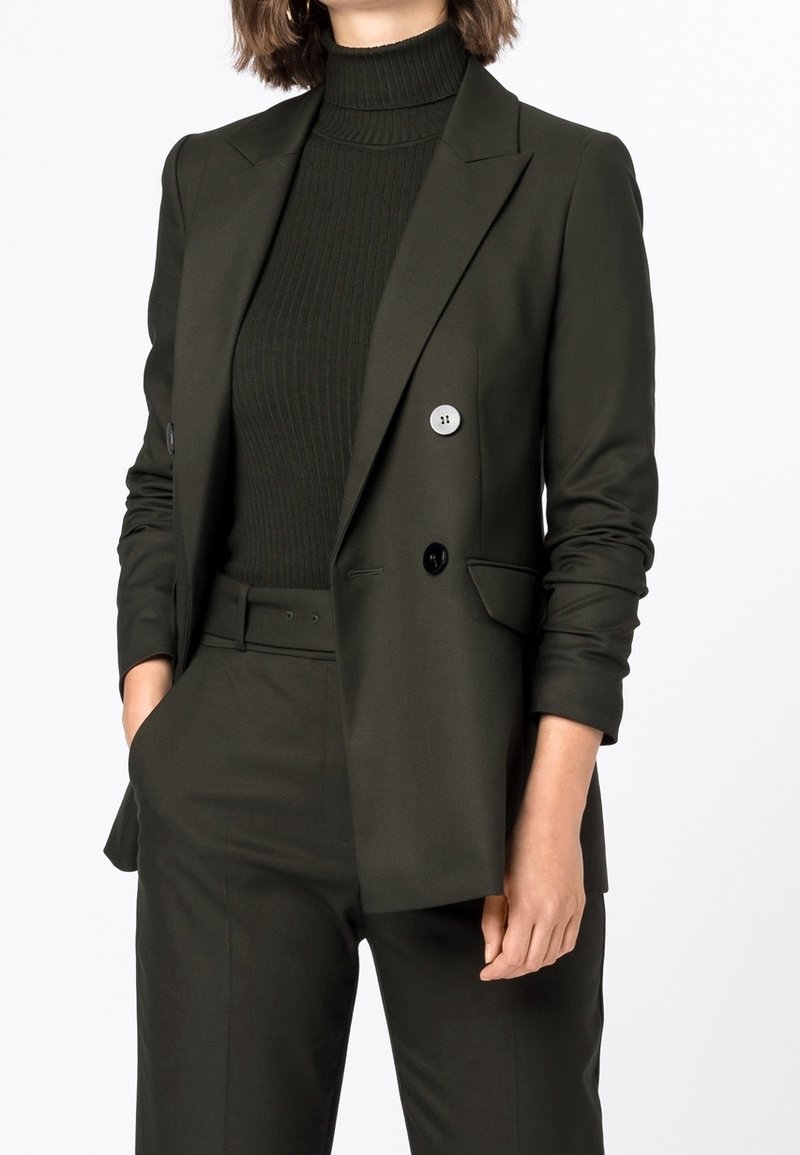 Tmavě zelený upravený blazer s dvojitými knoflíky, zkombinovaný s riflovým rolákem. Hladká látka, strukturované ramena a přiléhavý silueta.