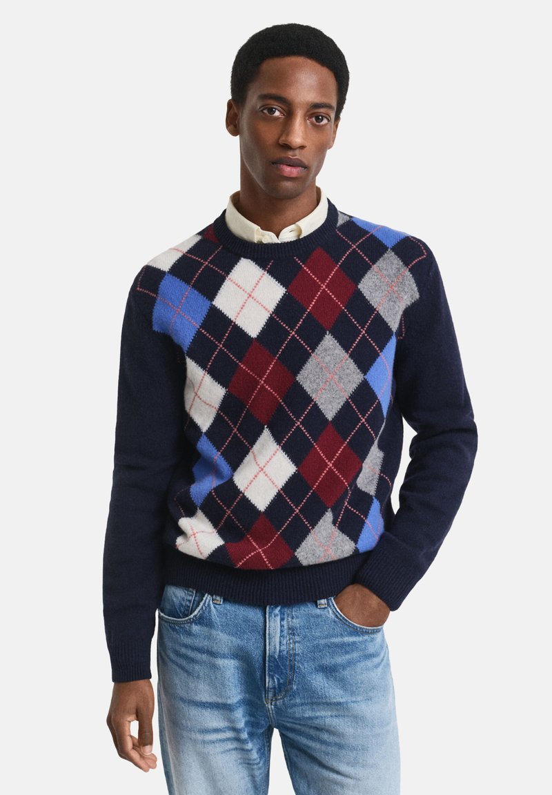 Marineblauer Argyle-Pullover mit roten, grauen und blauen Rautenmustern, gerippten Bündchen und Saum, aus weichem Material. Getragen über einem Hemd mit Kragen.