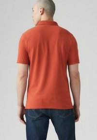 Kortärmad polo t-shirt i bränd orange bomull, med klassisk krage, jämn struktur och rak fåll, som bärs med mörkblå jeans.