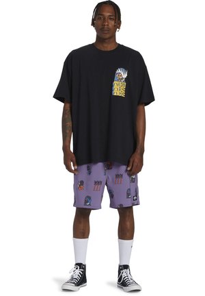 T-shirt nera oversize con una grafica colorata sul petto, abbinata a pantaloni corti viola con vivaci motivi tropicali e di personaggi.
