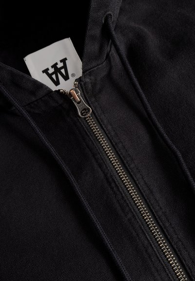 Sweat-shirt à capuche noir avec une fermeture éclair lisse et une étiquette arborant un logo. Le matériau semble texturé et durable, avec des détails de coutures visibles.