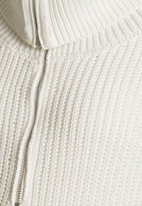 Suéter branco de malha com colar alto e fecho frontal. Apresenta um padrão texturizado com um toque robusto e acolhedor.